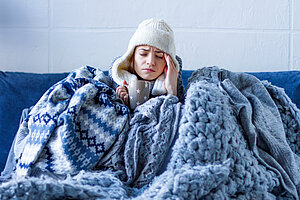 Frau sitzt erkältet und mit Mütze und Decke auf dem Sofa